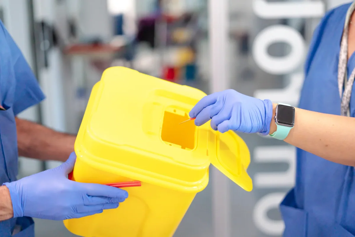 Gloved clinical staff handling a yellow sharps container.