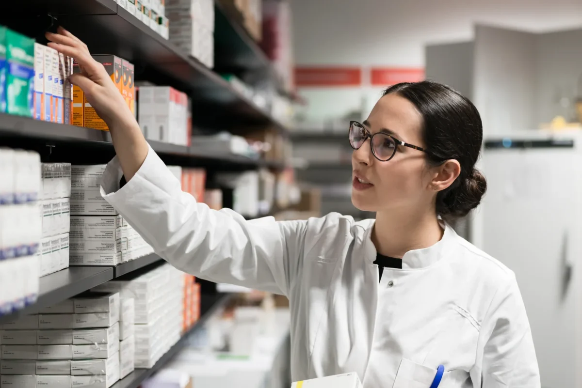 Pharmacist checking packaged medicines on pharmacy shelving.